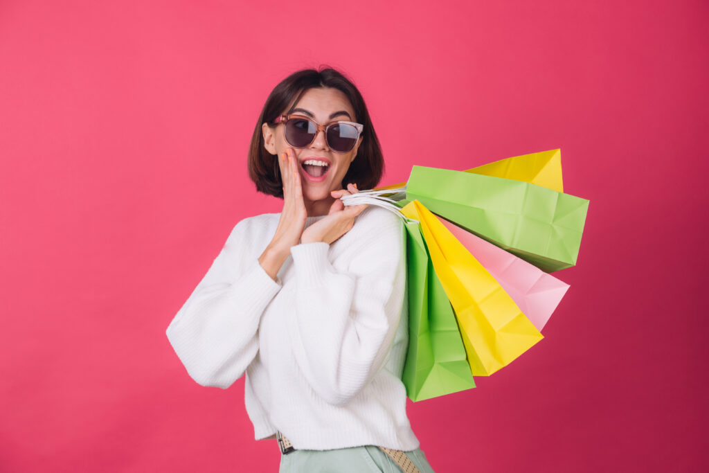 Reasons to Choose Many Parcel for Your Daily Grocery Needs woman in casual white sweater and sunglasses on pink red background holding shopping bags shocked amazed excited isolated copy space